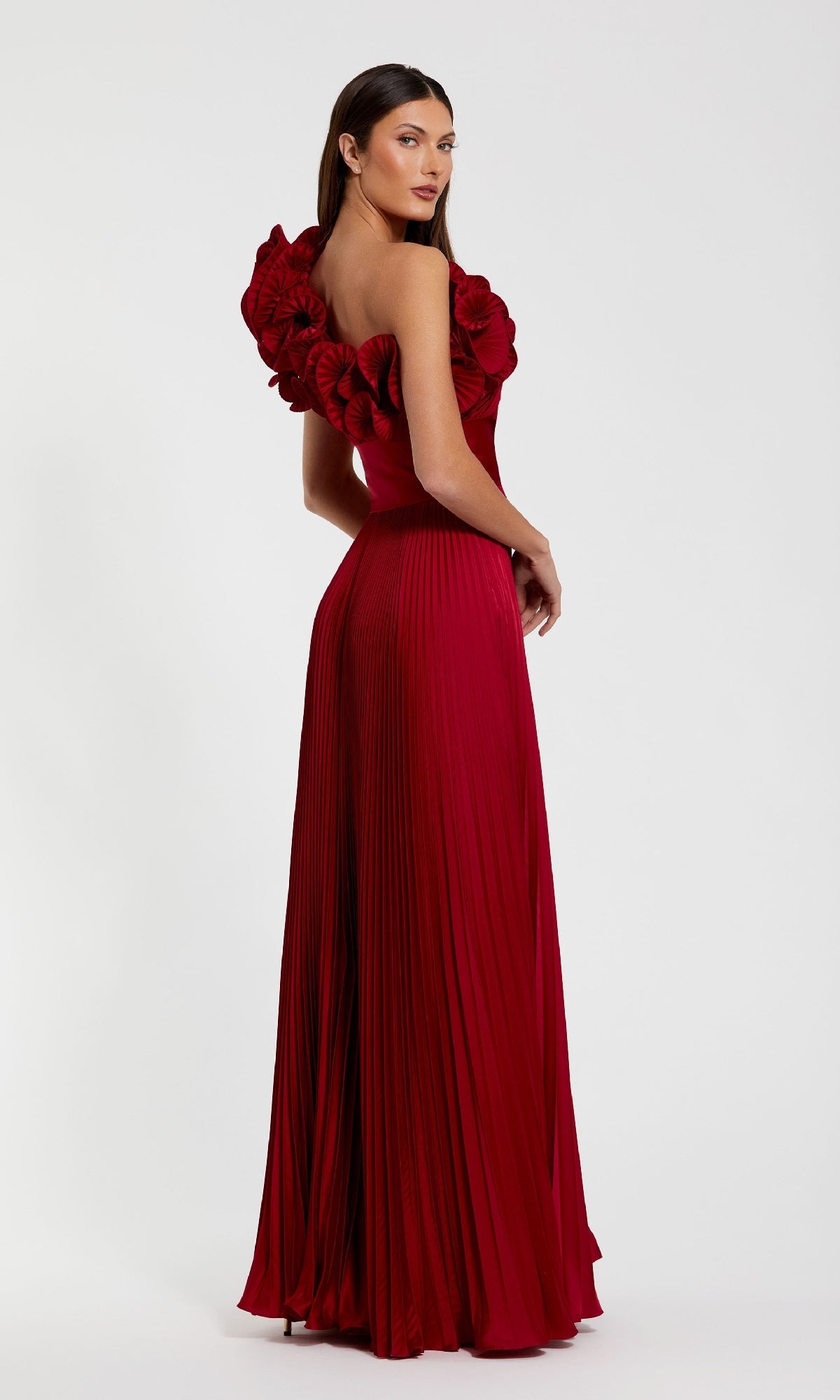 A woman models the Mac Duggal 49902 Pleated One-Shoulder Prom Gown, a floor-length, deep red dress featuring large floral details on one shoulder, against a plain white background.